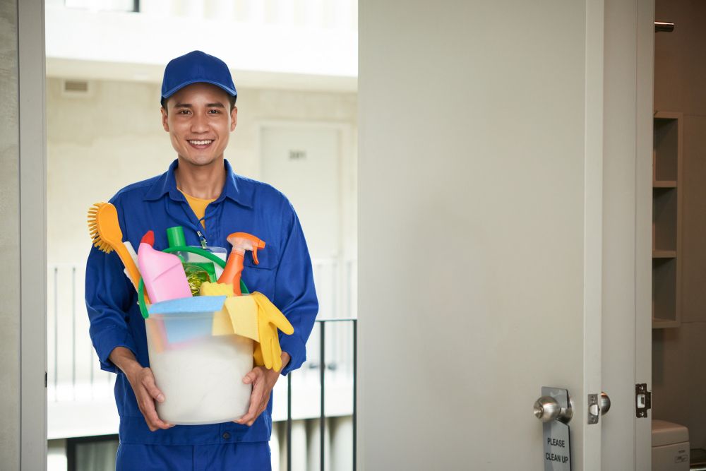 cheerful-asian-male-janitor-walking-into-hotel-room-carrying-supplies-in-bucket