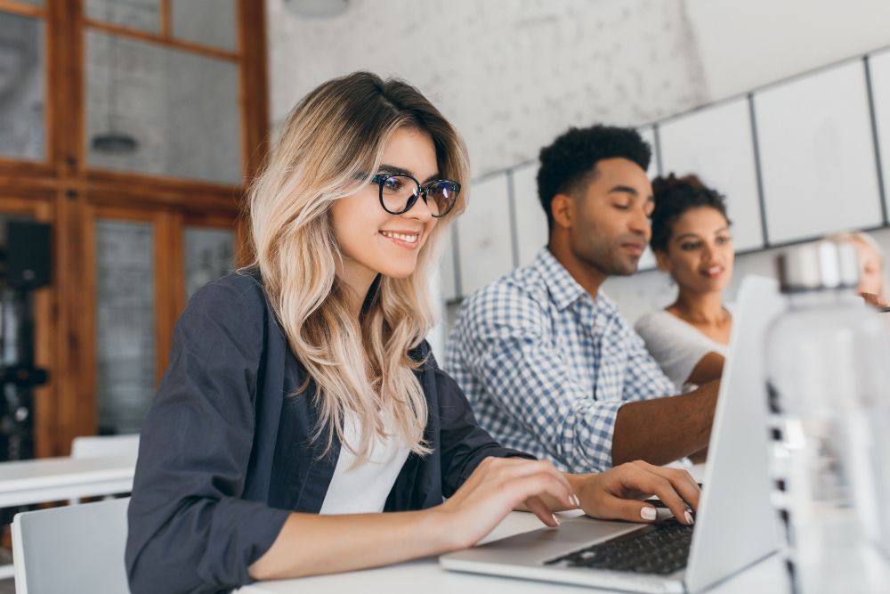 beautiful-curly-female-freelancer-with-cute-manicure-using-laptop-and-smiling-indoor-portrait-of-blonde-secretary-sitting-beside-african-coworker-in-blue-shirt