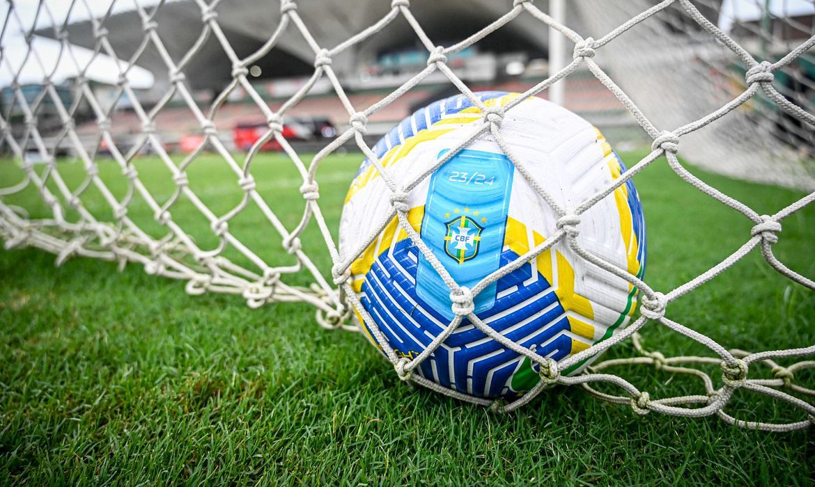 Bola oficial da CBF presa na rede do gol, com gramado e arquibancada ao fundo