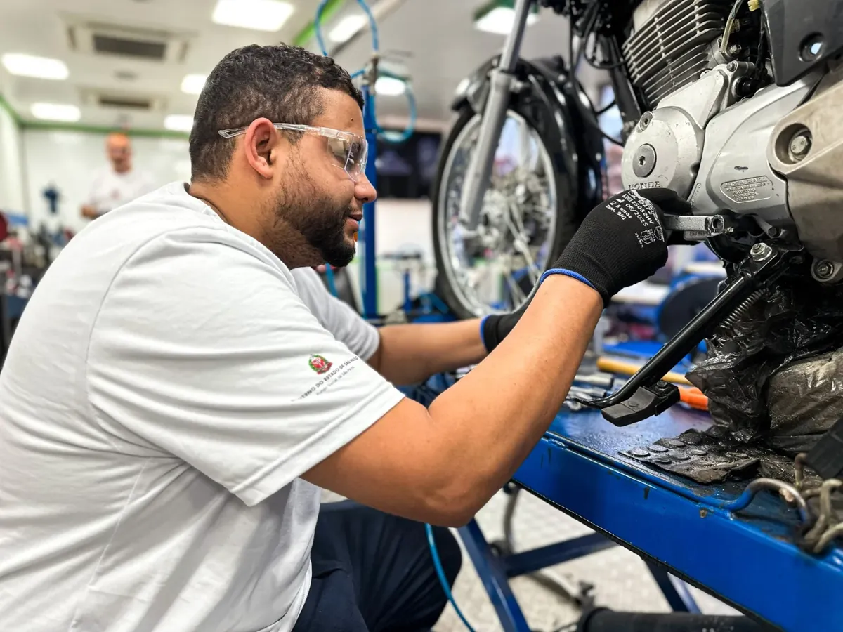 Homem usa óculos de proteção e luva enquanto conserta motor de motocicleta em oficina, vestindo camiseta com brasão do Governo do Estado de São Paulo