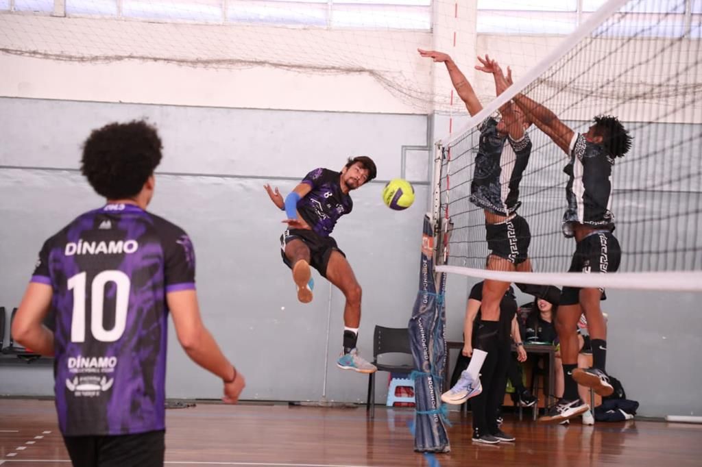Jogador salta para atacar a bola durante partida de vôlei, com adversários bloqueando no lado oposto da rede, em quadra do Campeonato Amador de Vôlei de Barueri.