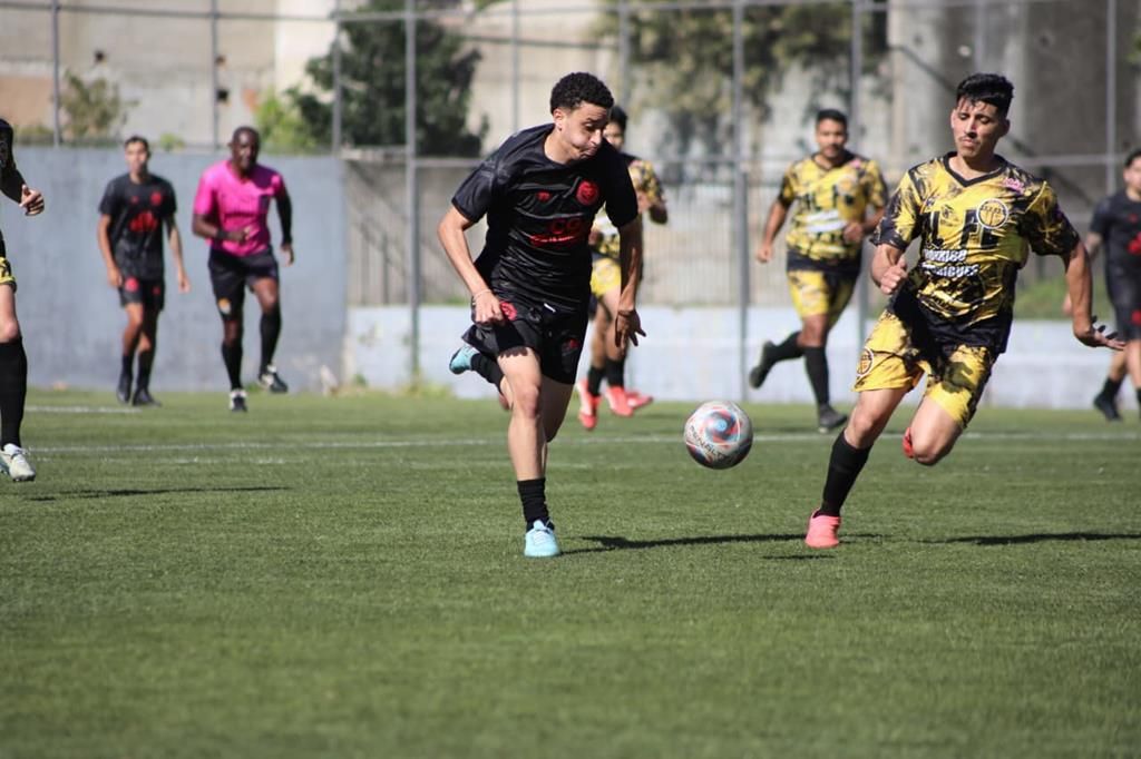 Jogadores de times de camisa preta e amarela disputam a bola durante partida do Campeonato Amador de Barueri, com árbitro de camisa rosa ao fundo