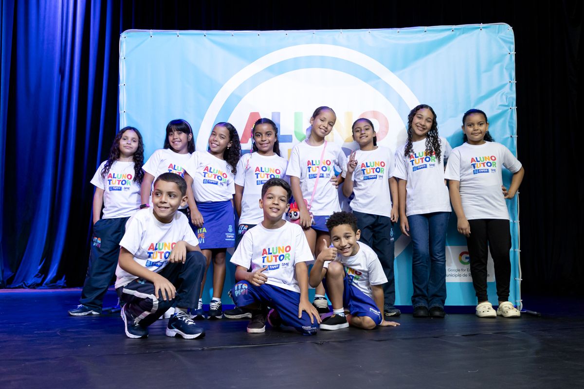 Grupo de crianças com camisetas brancas escritas Aluno Tutor posa em palco diante de painel azul do programa e do logotipo do Google for Education