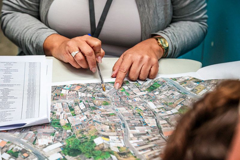 Mãos de uma técnica seguram um lápis sobre mapa aéreo de bairro, ao lado de lista de imóveis, durante trabalho de regularização fundiária em Itapevi
