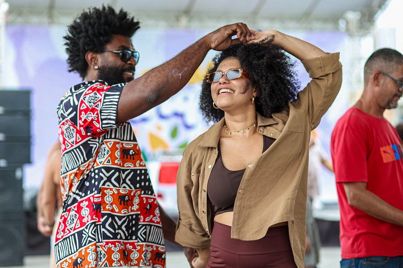 Casal dança samba rock sorrindo em evento ao ar livre; o homem gira a parceira pelo braço enquanto outras pessoas dançam ao fundo, em estrutura de palco coberta.